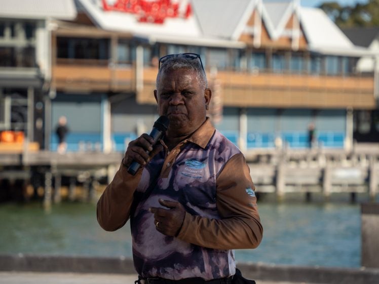 Image of An older man wearing sunglasses on his head speaks into a microphone outdoors, standing near water with blurred buildings in the background.