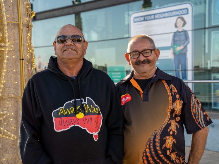 Image of Two men standing and smiling outdoors; one wears sunglasses and a black Always Was Always Will Be hoodie, the other wears glasses and a black shirt with orange Indigenous designs. There are lights and a building in the background.