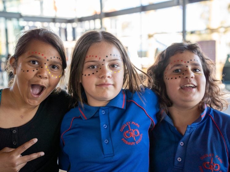 Image of Three children with face paint smile and pose together indoors. Two wear blue Quigley Park Primary School uniforms, while the third wears black and makes a peace sign. Sunlight filters in through large windows behind them.