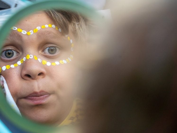 Image of A child with painted yellow and white dots around their eyes looks at their reflection in a round mirror, their expression curious and focused.
