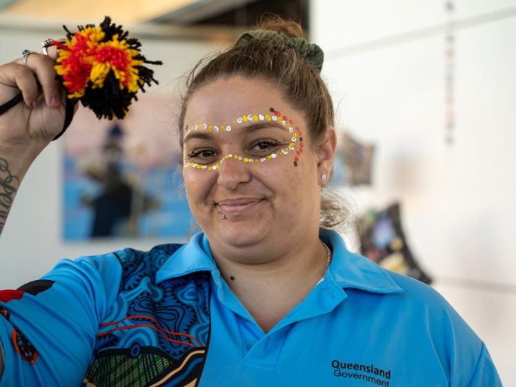 Image of A woman wearing a blue Queensland Government shirt and traditional face paint smiles whilst holding up a handmade craft with red, yellow, and black wool. Indigenous artwork decorates part of her shirt.