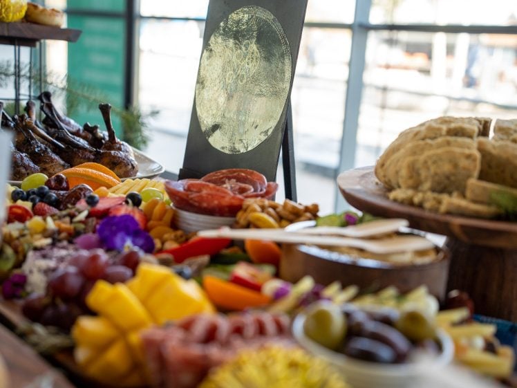 Image of A colourful charcuterie table with assorted cheeses, cured meats, fruits, bread, nuts, and dips. A decorative sign stands in the background, and sunlight streams through the window behind the spread.
