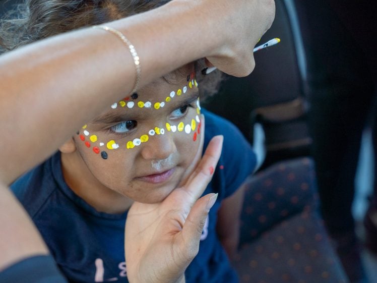 Image of A child is getting their face painted with rows of yellow, white, and red dots. An adult’s hand gently holds the child’s chin and paints near the child’s eyes. The child is looking to the side.