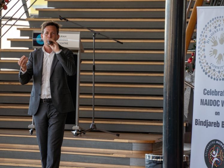 Image of A man in a suit speaks into a microphone whilst standing in front of stairs. A sign nearby mentions NAIDOC Week and the Bindjareb community. A podium and microphone stand are visible in the background.
