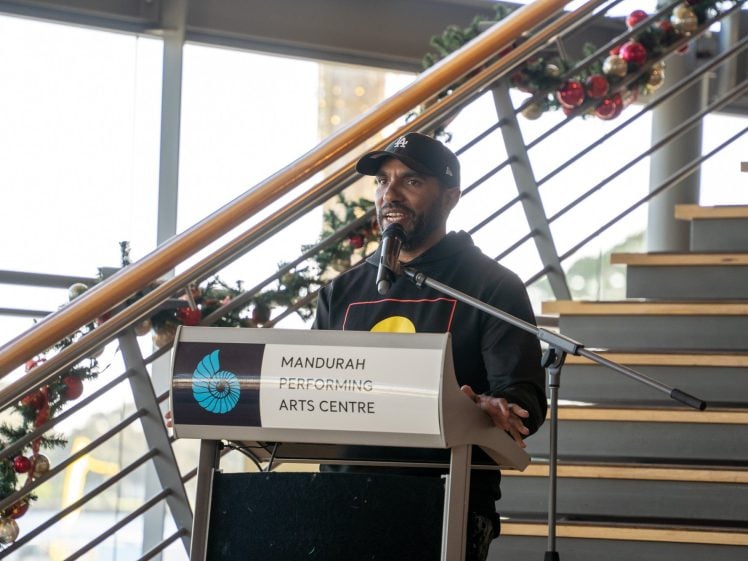 Image of A man speaks into a microphone at a lectern labelled Mandurah Performing Arts Centre, with festive garland wrapped around a staircase in the background.