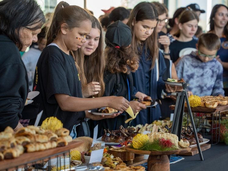 Image of A group of young people, some with painted faces, serve themselves food from a buffet table filled with pastries and savoury dishes at a crowded indoor event.