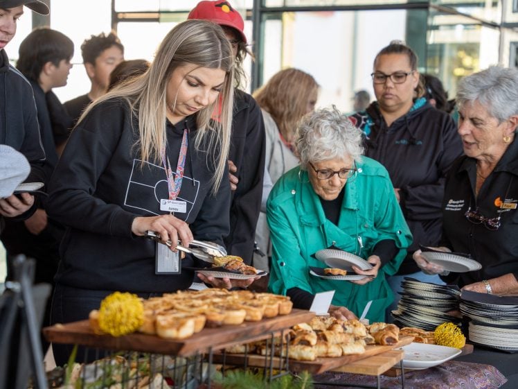 Image of A group of people gather around a table filled with pastries and food. A woman in a black hoodie serves food with tongs whilst others wait, holding plates and watching. The scene appears to be at a casual event or gathering.