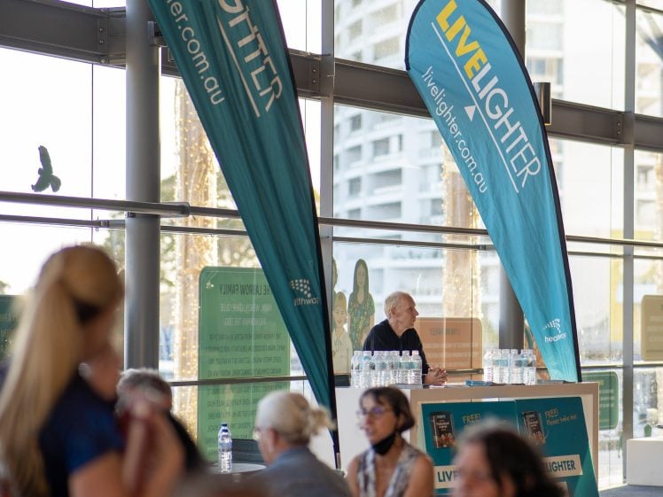 Image of People gather indoors near tall blue LiveLighter banners and a stand with health pamphlets and water bottles. A person stands behind the stand, and city buildings are visible through large windows in the background.