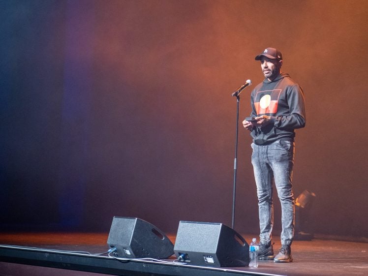 Image of A person wearing a hoodie, jeans, and a cap stands on stage speaking into a microphone. There are two speakers and a water bottle on the stage, with orange and blue lighting in the background.
