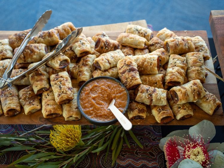 Image of A wooden platter filled with golden-brown pastry rolls, sprinkled with seeds, is served with a bowl of orange dipping sauce and tongs. The platter is decorated with native Australian flowers and greenery.