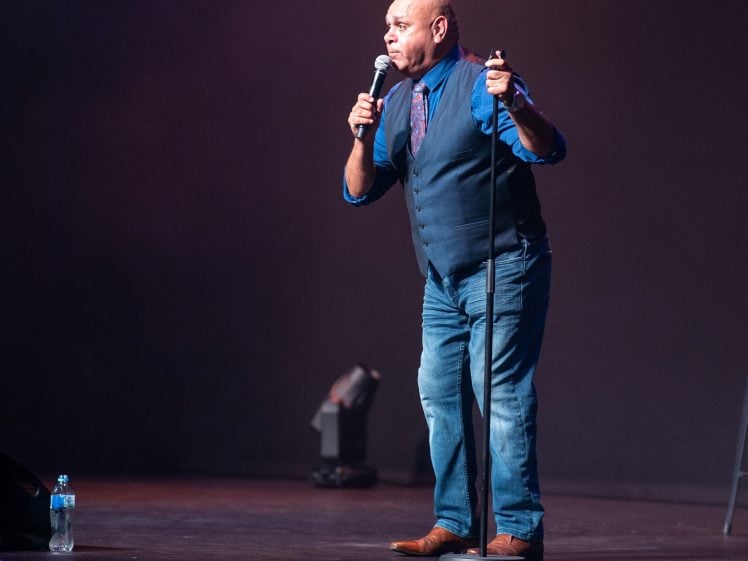 Image of A man in a blue shirt, jeans, and a waistcoat performs stand-up comedy on stage, holding a microphone and stand. A water bottle sits on the floor nearby under stage lighting.
