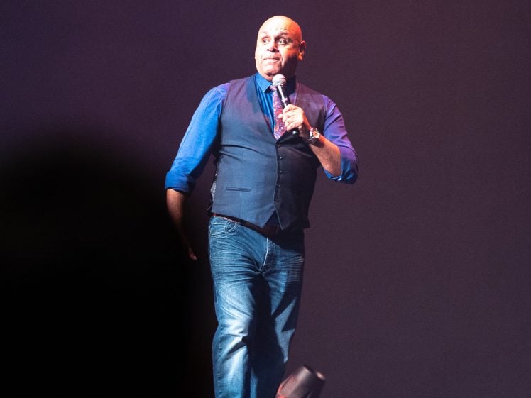 Image of A man in a blue shirt and waistcoat stands on stage holding a microphone, appearing to speak or perform. The background is dark and he is lit by stage lights.