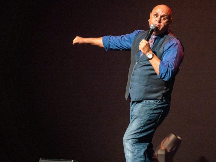 Image of A bald man in a blue shirt, waistcoat, and jeans holds a microphone and gestures energetically on stage during a performance.