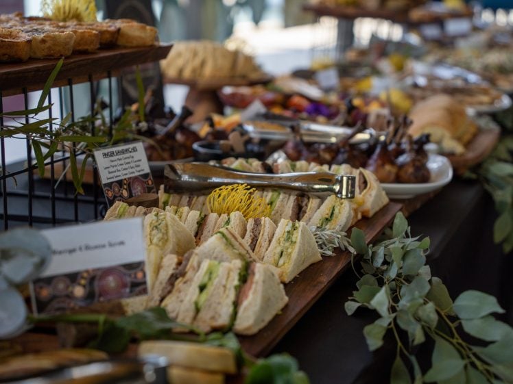 Image of A buffet table displays an assortment of food, including assorted sandwiches, pastries, and appetisers, arranged on wooden trays with decorative greenery and serving tongs.