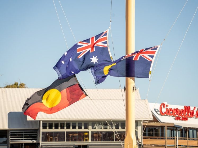 Image of Four flags fly on a pole: the Australian flag, the Torres Strait Islander flag, the Aboriginal flag, and another flag. In the background, there’s a building with a red Cicerello’s sign. The sky is clear and blue.