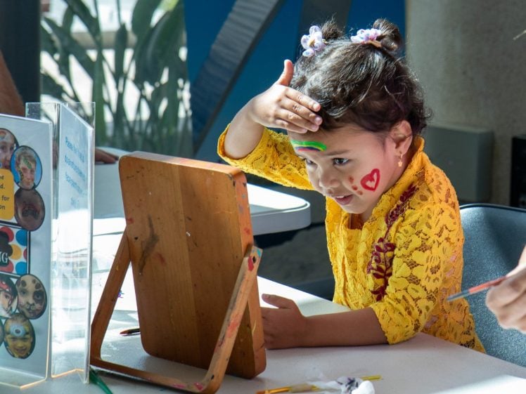 Image of A young girl in a yellow dress sits at a table, looking into a mirror while touching her forehead. Her face is painted with a red heart and green lines, and she appears to be enjoying a face-painting activity.