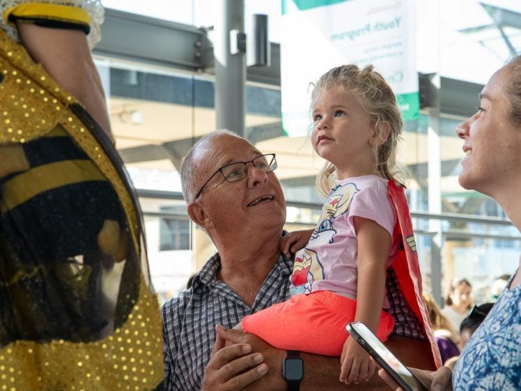 Image of An older man holds a young girl in bright clothes as they both look up and smile at a person in a yellow costume. A woman stands beside them, holding a mobile and smiling. They are indoors with sunlight streaming in.