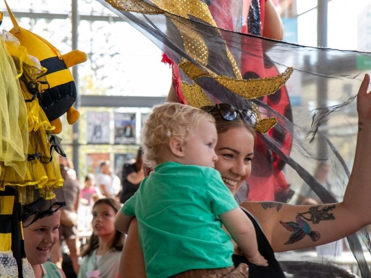 Image of A smiling woman holding a small child lifts a sheer black fabric decorated with gold. Children in fancy dress, including a bee outfit, watch nearby in a busy, sunlit indoor setting.