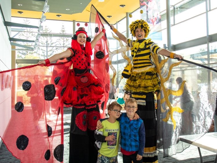 Image of Two adults on stilts dressed as a ladybird and a bee pose indoors with two smiling children. The ladybird holds sheer red wings with black spots, while the bee has yellow and black striped wings. Sunlight streams through large windows.