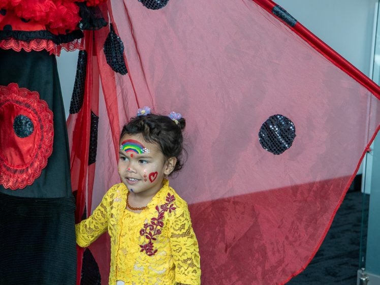 Image of A young child with face paint, including a rainbow and hearts, stands smiling beneath large red and black ladybird wings. The child wears a bright yellow lace top and is indoors next to someone dressed in a matching ladybird-themed costume.