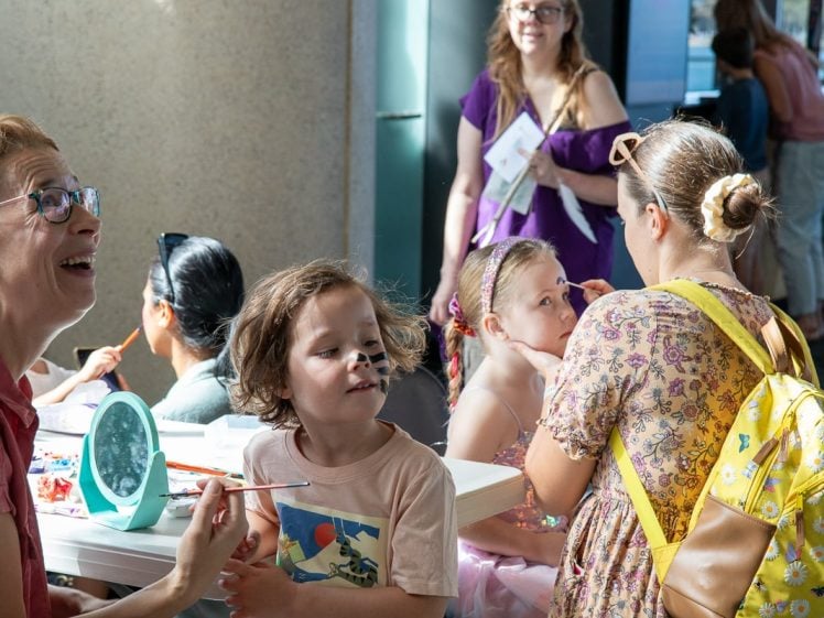 Image of Children and adults gather at a table for face painting. A woman smiles whilst painting a child’s face; another child wears a sparkly dress and headband. People in casual clothing stand and watch in a brightly lit indoor space.