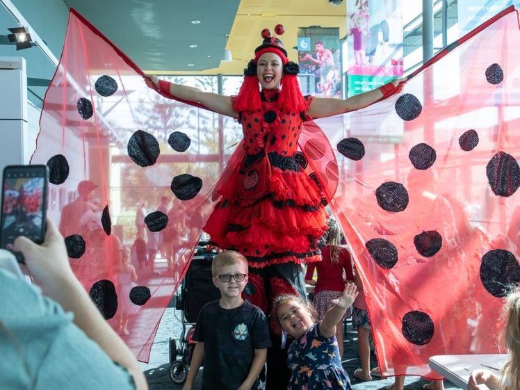 Image of A performer in a red ladybird costume with large wings poses with two smiling children in front. People around them take photos in a brightly lit indoor space with colourful decorations.