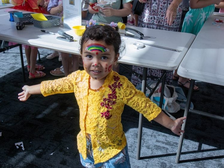 Image of A young child wearing a yellow lace top with face paint, including a rainbow and hearts, stands with arms open, smiling in a brightly lit room with tables and children in the background.