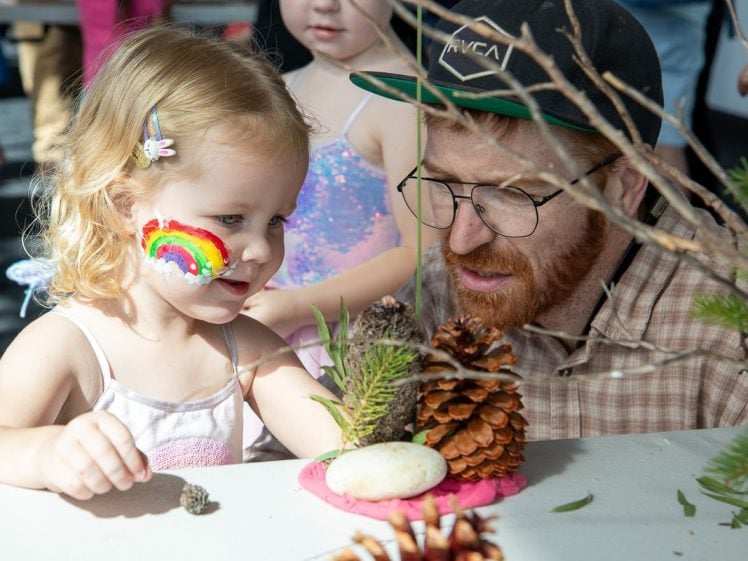 Image of A young girl with a rainbow painted on her cheek and a man in glasses and a cap create nature crafts with pine cones, rocks, and clay at a table. Other children are visible in the background.