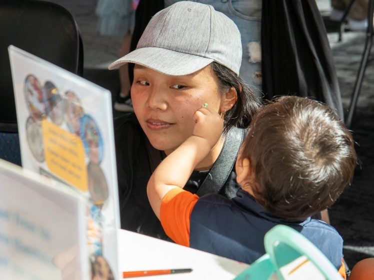Image of A young child touches the cheek of an adult wearing a grey hat, who is sitting at a table with leaflets and a pen in a brightly lit indoor setting.