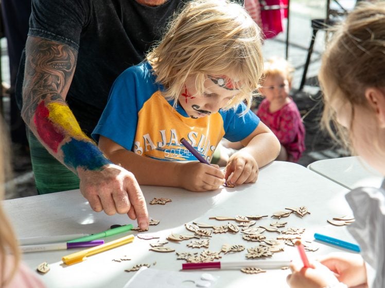 Image of A child with face paint colours a wooden shape at a table covered with craft supplies. An adult with a colourful painted arm assists, while other children participate in the background. Sunlight streams in, creating a cheerful scene.