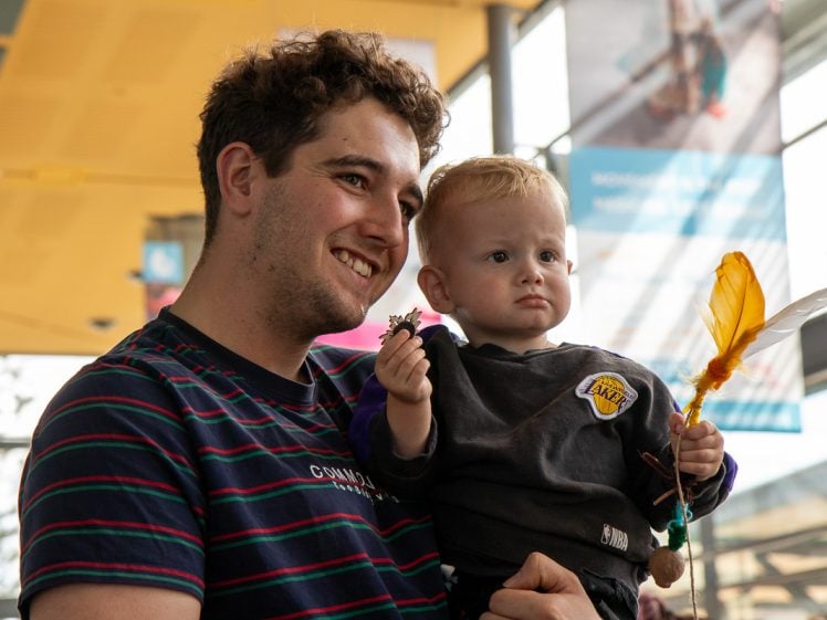 Image of A man in a striped shirt smiles whilst holding a toddler. The toddler, wearing a Lakers shirt, holds a small feathered object and looks serious. They are indoors, with blurred posters and bright windows in the background.