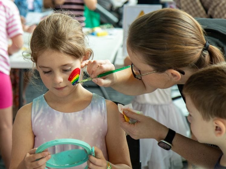 Image of A woman paints a rainbow on a young girl's face at a table, while the girl holds a mirror. A boy watches nearby. The setting appears to be a lively indoor event with other children in the background.