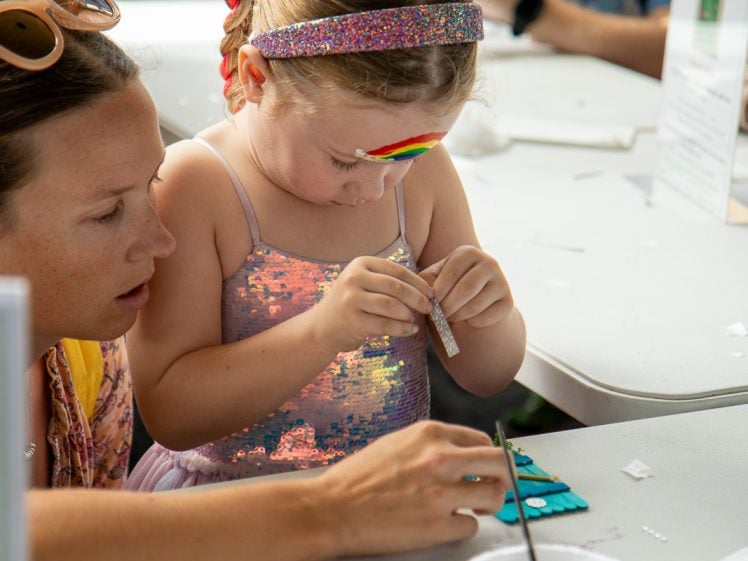 Image of A young girl with a rainbow painted on her forehead, wearing a glittery dress and headband, concentrates on a craft project at a table, assisted by an adult woman beside her. Craft materials are scattered on the table.