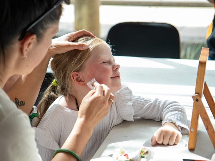 Image of A woman cleans a young girl’s face with a tissue while the girl smiles and sits at a table, eyes closed, with sunlight streaming in. A small mirror and face painting supplies are on the table.