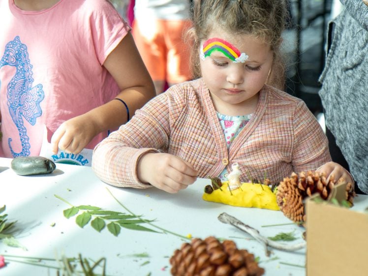 Image of A young child with a rainbow painted on their face makes crafts with natural materials like leaves and pine cones at a table, surrounded by other children.
