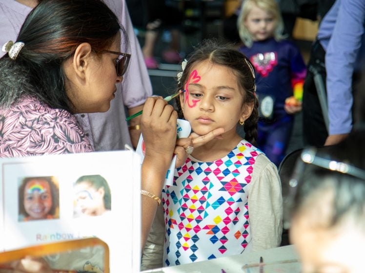 Image of A woman paints a red butterfly on a young girl's face at a face painting stall. The girl stands still, wearing a colourful geometric-patterned dress. Other children and adults are visible in the background.