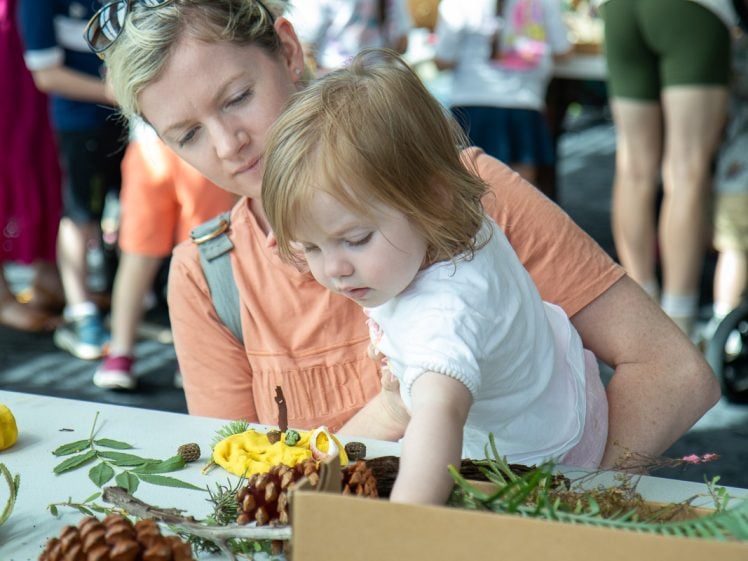 Image of A woman helps a young child reach for natural craft materials, such as pine cones and leaves, on a table at a busy indoor event. Other people and craft supplies are visible in the background.