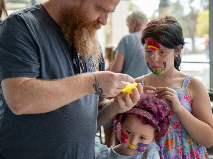 Image of A bearded man helps two children with face paint; one child has colourful paint on her face and a wig, the other wears a dress and watches closely. They are indoors, with light coming through large windows.