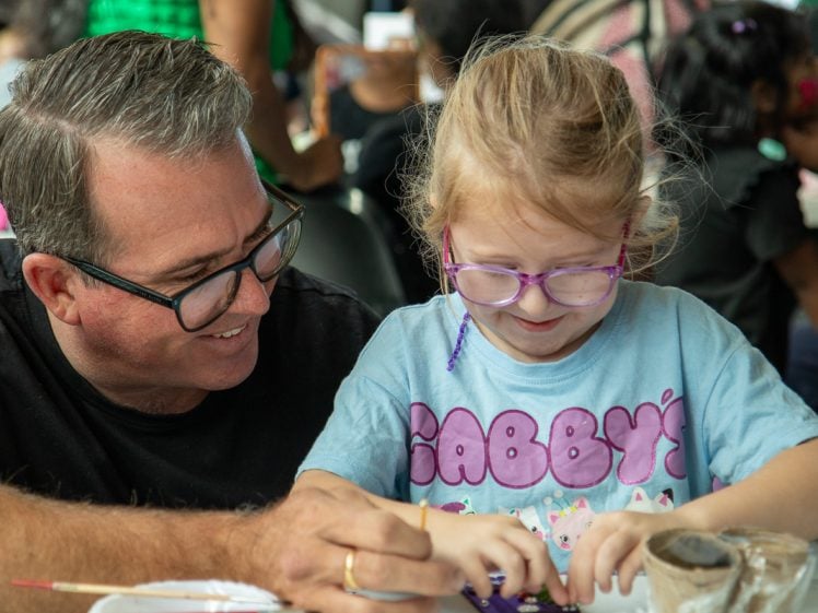 Image of A man and a young girl with glasses sit together at a table, smiling and working on a craft project. The girl wears a blue shirt that says Gabbys. Other people are visible in the background.