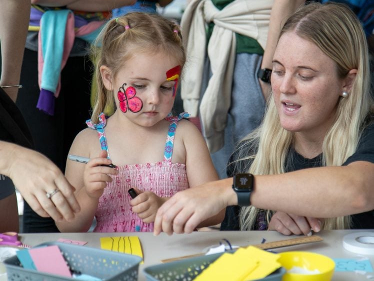 Image of A young girl with a butterfly painted on her face sits at a table doing crafts with a woman. The woman is guiding her as they work with colourful paper and craft supplies. Other people are visible in the background.