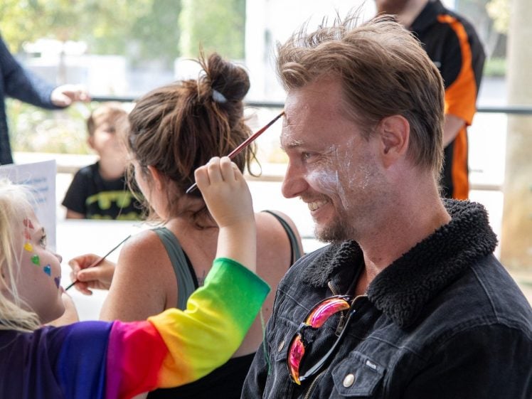 Image of A smiling man sits while a child in a rainbow-coloured shirt paints his face. The man has white paint on his forehead and is wearing a black jacket with sunglasses hanging from the collar. Other people are in the background.