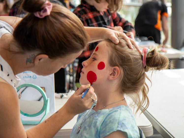 Image of A woman paints large red dots on a young girl's face at a table, whilst other children are visible in the background, suggesting a face painting activity or event.