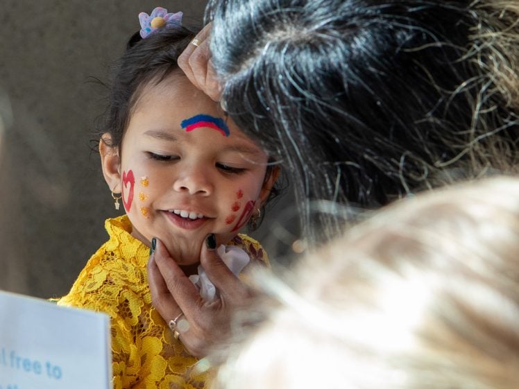Image of A young girl in a yellow dress smiles as someone paints her face. She has colourful designs on her cheeks and forehead, and light shines on her from the side. Others are nearby, partially visible in the frame.