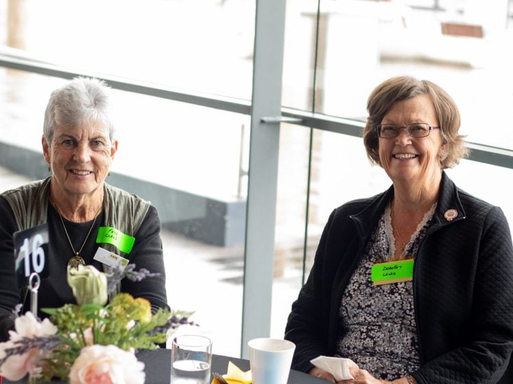Image of Two older women wearing glasses and name badges sit at a table with flowers, drinks, and a table number 16 in a bright, modern indoor setting with large windows. They are smiling at the camera.