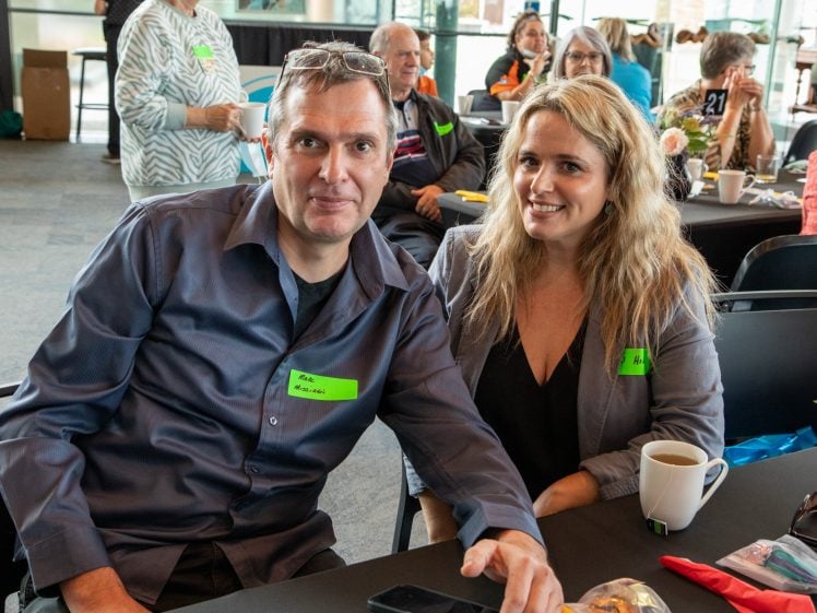 Image of A man and a woman sit next to each other at a table during an indoor event. Both wear name badges and smile at the camera. Other people are seated at tables in the background. A coffee cup is on the table.