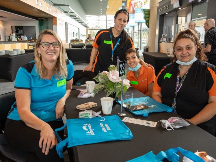 Image of Four smiling women, wearing blue and orange shirts, sit around a table with blue bags, cups, and flowers in a bright, modern indoor setting.