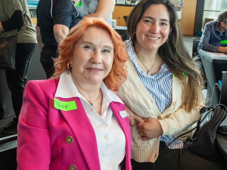 Image of Two women smiling at an indoor event. One has red hair, a pink blazer, and a name badge reading Trish. The other has long brown hair and is wearing a cream jumper over a blue striped shirt. People are in the background.