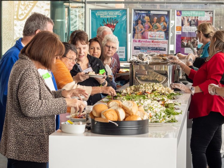 Image of A group of people serve themselves food from a buffet table with salads, bread, and hot dishes. Several servers wearing gloves assist behind the counter. Posters and large windows are visible in the background.