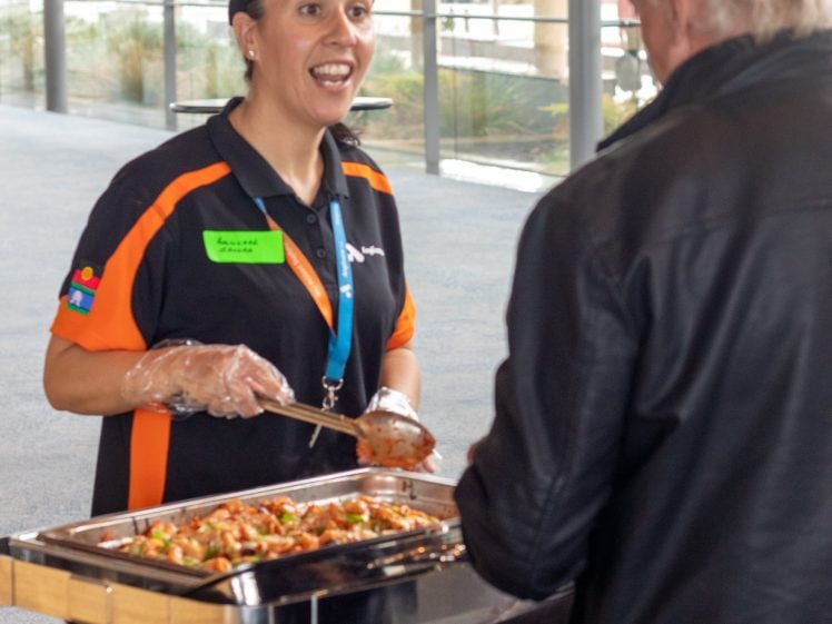 Image of A woman wearing gloves and a black-and-orange uniform serves food from a chafing dish to an older man in a black jacket at an indoor event.