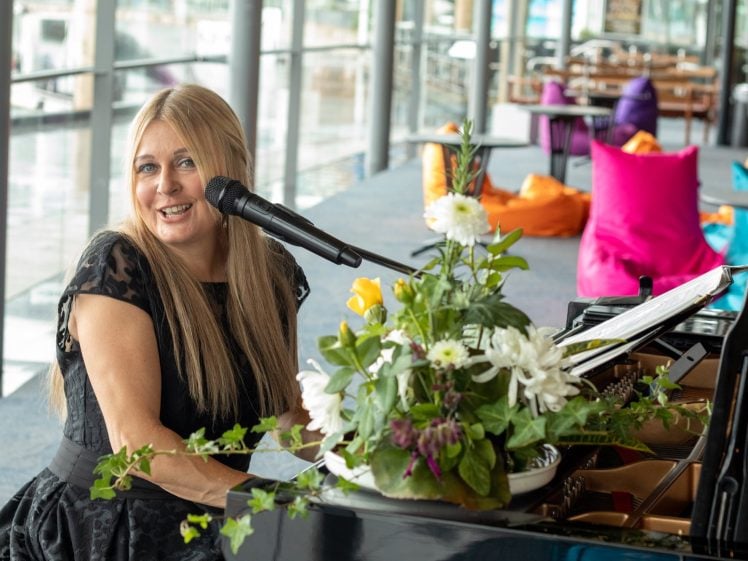 Image of A woman with long blonde hair smiles whilst playing the piano and singing into a microphone. A vase of flowers sits on the piano, and colourful beanbags are in the background by large windows.